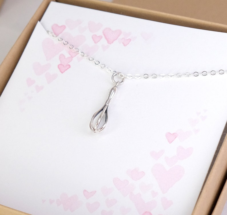 Puede incluir: Un collar de plata con un dije de batidor. El collar est&aacute; en una caja con un fondo rosa y blanco con corazones.