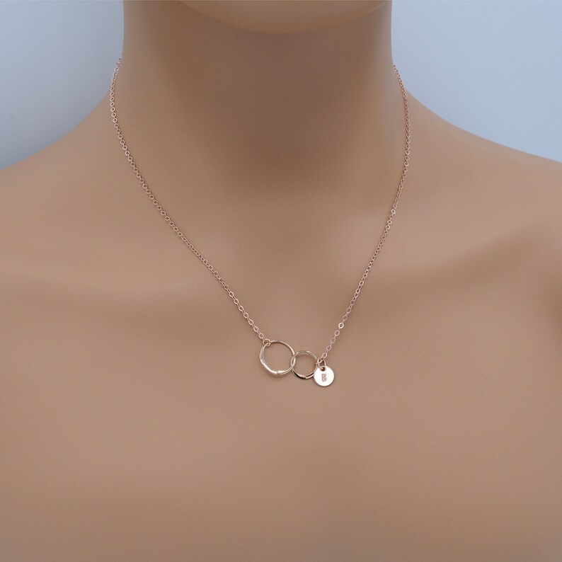 Pode incluir: Um colar de ouro rosa com dois c&iacute;rculos entrela&ccedil;ados e um pequeno pingente redondo com a letra 'R' gravada.