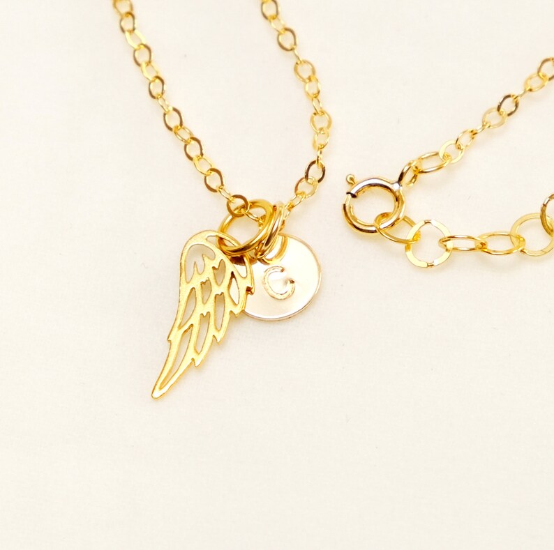 May include: Gold chain necklace with a small gold angel wing charm and a gold disc charm with the letter 'G' engraved on it.