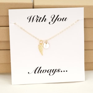 May include: A gold necklace with a small angel wing charm and a round charm with the letter "S" on it. The necklace is displayed on a white card with the text "With You Always..."