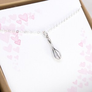 Puede incluir: Un collar de plata con un peque&ntilde;o dije de batidor. El collar est&aacute; sobre un fondo blanco con corazones rosados de acuarela.