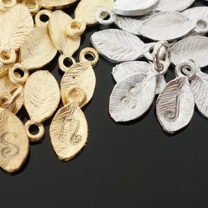 May include: A collection of gold and silver leaf charms with engraved letters. The charms are shaped like leaves and have a textured surface.