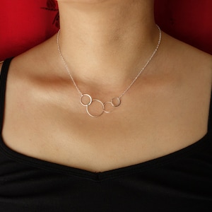 May include: A silver necklace featuring five interconnected circles of varying sizes. The circles are suspended from a delicate chain. The necklace is displayed against a black top and a red background.
