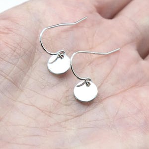 May include: A pair of silver-toned drop earrings featuring small, circular, flat metal discs suspended from curved ear wires. The earrings have a polished, reflective surface and a simple, clean design that hangs vertically from the hook mechanism.