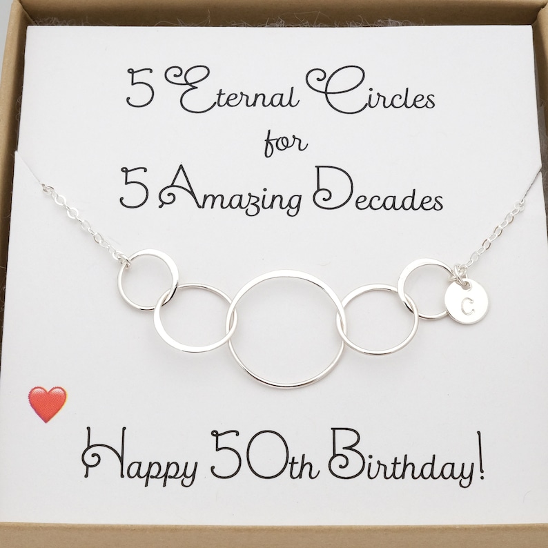 K&ouml;nnte beinhalten: Eine silberne Halskette mit f&uuml;nf ineinandergreifenden Kreisen und einem Anh&auml;nger mit dem Buchstaben "C". Die Halskette befindet sich in einer Geschenkbox mit einer Karte, auf der steht: "5 Ewige Kreise f&uuml;r 5 Erstaunliche Jahrzehnte Herzlichen Gl&uuml;ckwunsch zum 50. Geburtstag!"