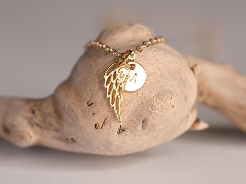 May include: Gold necklace with a delicate angel wing charm and a small round charm with the letter "M" engraved on it.