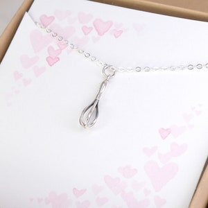 Puede incluir: Un collar de plata con un peque&ntilde;o dije de batidor. El collar est&aacute; sobre un fondo blanco con corazones rosas.