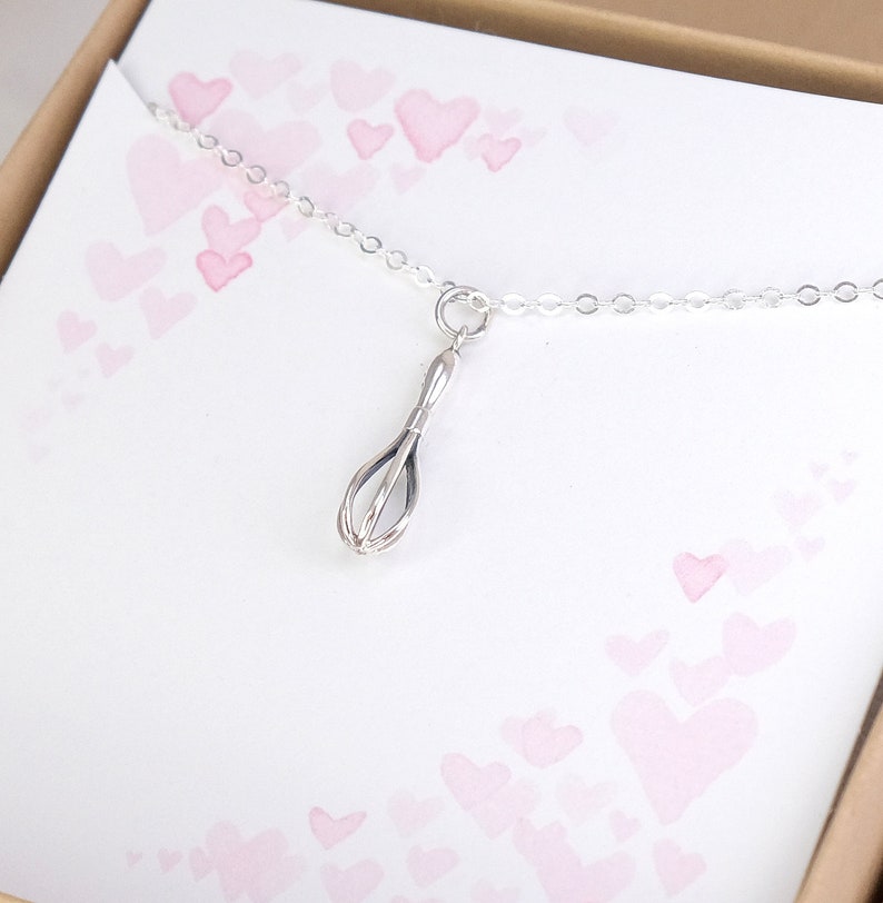 Puede incluir: Un collar de plata con un peque&ntilde;o dije de batidor. El collar est&aacute; sobre un fondo blanco con corazones rosas.
