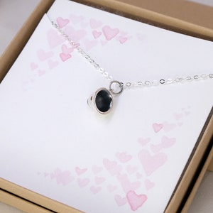 May include: A silver necklace with a small, round, black pendant. The pendant has a textured surface and is attached to a delicate silver chain. The necklace is displayed in a box with a pink and white heart pattern.