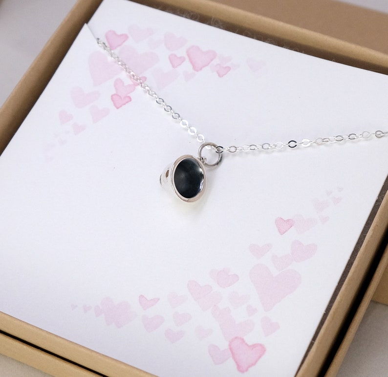 May include: A silver necklace with a small, round, black pendant. The pendant has a textured surface and is attached to a delicate silver chain. The necklace is displayed in a box with a pink and white heart pattern.