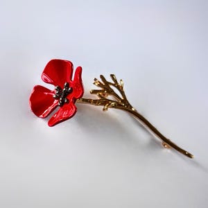 May include: A red poppy flower brooch with a gold-coloured stem and branch detail. The brooch features a bright red enamel flower with a dark centre. The gold stem extends to the right, creating an elegant floral accessory.