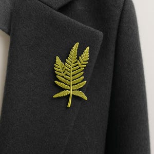 May include: A gold-colored fern leaf brooch pinned to a dark gray blazer. The brooch is a detailed representation of a fern frond, with intricate veining and a textured surface. The blazer's fabric has a subtle, felted appearance.