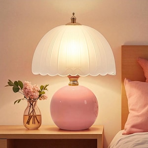 May include: A pink ceramic table lamp with a white, scalloped lampshade. The lamp has a round base and a gold-coloured accent at the base of the shade. A small vase of flowers sits next to the lamp on a wooden surface.