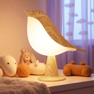 Könnte beinhalten: Ein Nachtlicht in Form eines Vogels aus Holz mit einem hellweißen, leuchtenden Körper. Die Lampe hat eine helle Holzmaserung und steht auf einem passenden Holzsockel. Kuscheltiere sind im Hintergrund.