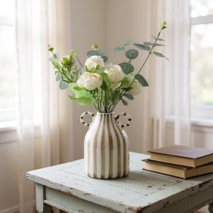 Puede incluir: Un jarrón de cerámica a rayas blancas y beige con dos asas contiene un ramo de flores blancas y follaje verde. El jarrón está sobre una mesa de madera azul claro desgastada junto a dos libros apilados con tapas marrones.