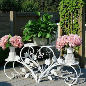 Può includere: Un portapiante bianco e ornato a tre livelli con un design floreale. Il supporto contiene ortensie rosa in vasi bianchi e una pianta verde in un vaso grigio. Il supporto è in metallo e si trova su un terrazzo di legno.