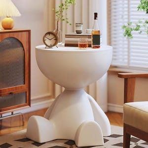 Puede incluir: Una mesa auxiliar blanca y moderna con un diseño único que se asemeja a una figura sentada. La mesa tiene una parte superior e inferior redondeadas, con un soporte central. Un pequeño reloj, libros y un vaso de líquido ámbar están sobre la mesa. La mesa está en un salón.