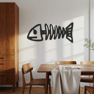 以下が含まれることがあります： 黒い金属製の魚の骨格の壁の装飾。このアートワークは、木製のダイニングテーブルの上にある白い壁に取り付けられています。魚の骨格のデザインは、頭、背骨、尾を特徴とし、ミニマリストでモダンな美学を作り出しています。