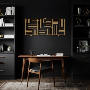 Op de afbeelding: Een goudkleurig, geometrisch kunstwerk aan de muur met een herhalend patroon van rechthoekige vormen hangt boven een houten bureau. Het bureau staat in een thuiskantoor met zwarte boekenkasten en een houten stoel.