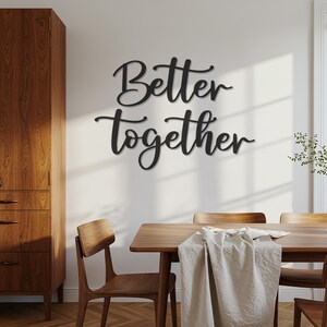 May include: Black metal wall art spelling "Better Together" in a cursive font. The sign is mounted on a white wall above a wooden dining table and chairs. A linen tablecloth is draped across the table.