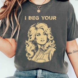 Könnte beinhalten: Ein anthrazitfarbenes Kurzarm-T-Shirt mit einem gelben Grafikdruck einer Frau mit voluminösem Haar und Creolen. Der Text "I BEG YOUR" ist in Gelb über der Illustration aufgedruckt. Das T-Shirt hat einen Rundhalsausschnitt und eine lockere Passform.