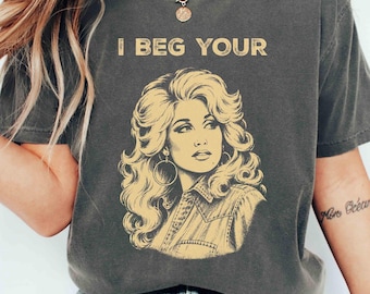 May include: A charcoal gray short-sleeve t-shirt featuring a yellow graphic print of a woman with voluminous hair and hoop earrings. The text "I BEG YOUR" is printed in yellow above the illustration. The shirt has a crew neckline and a relaxed fit.