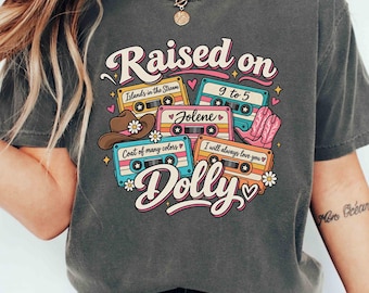 Könnte beinhalten: Ein anthrazitfarbenes T-Shirt mit einem Grafikdruck und dem Text Raised on Dolly. Das Design zeigt fünf bunte Kassetten mit den Beschriftungen Islands in the Stream, 9 to 5, Jolene, Coat of many colors und I will always love you sowie einen Cowboyhut und rosa Stiefel.
