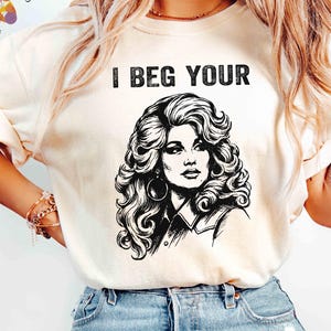 Könnte beinhalten: Ein cremefarbenes Kurzarm-T-Shirt mit einer schwarzen grafischen Illustration einer Person mit voluminösem, welligem Haar und großen Creolen. Über der Illustration steht der Text I BEG YOUR in einer fetten, schwarzen Schrift.