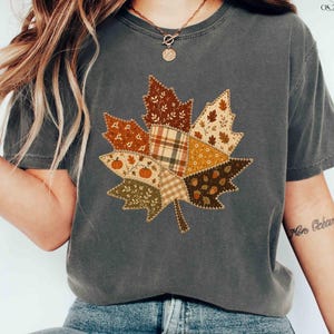 May include: A charcoal grey short-sleeve t-shirt featuring a large, central graphic of a maple leaf composed of various autumn-themed patchwork patterns, including plaid, floral, and pumpkin prints in shades of brown, orange, and cream.