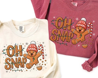 Könnte beinhalten: Zwei langärmlige Shirts, eines in Creme und eines in Weinrot, mit einem Lebkuchenmann-Motiv und dem Text "OH SNAP". Der Lebkuchenmann trägt einen Hut und hält eine Zuckerstange. Die Shirts sind mit Lichterketten und Schneeflocken verziert.