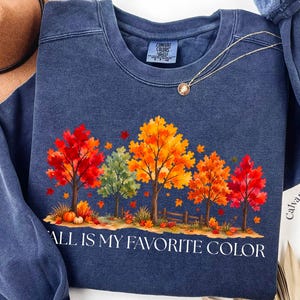 May include: A navy blue crewneck sweatshirt featuring a graphic of autumn trees with red, orange, and yellow leaves. The text "FALL IS MY FAVORITE COLOR" is printed in white below the trees. A small pumpkin sits near the base of the trees.