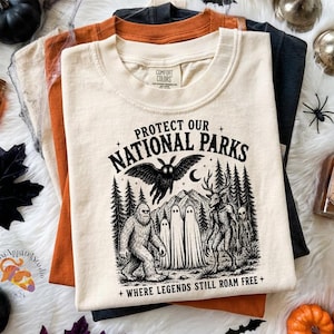 Op de afbeelding: Een crèmekleurig t-shirt met een zwarte grafische print met de tekst Protect Our National Parks Where Legends Still Roam Free. Het ontwerp toont een mot, Bigfoot, geesten en een hertachtig wezen in een bosrijke omgeving.