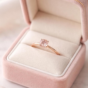 May include: A rose gold ring with a round, light pink gemstone, presented in a pink velvet ring box. The ring features a slender band and a simple, elegant design, suitable for an engagement or special event.