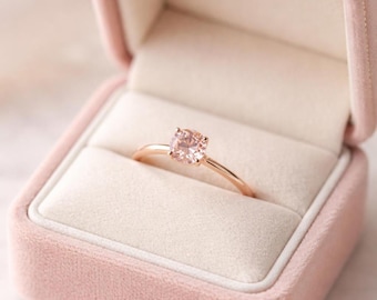 Op de afbeelding: Een roségouden ring met een ronde, lichtroze edelsteen, gepresenteerd in een roze fluwelen ringdoos. De ring heeft een slanke band en een eenvoudig, elegant ontwerp, ideaal voor een verloving of speciale gelegenheid.