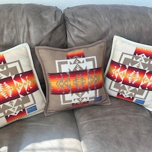 May include: Three square throw pillows with a geometric pattern in shades of red, orange, tan, and white. The pillows are arranged on a brown leather couch. Each pillow features a unique design with a central motif.