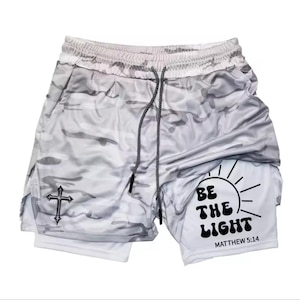 Puede incluir: Pantalones cortos deportivos de camuflaje blanco y gris con cordón negro. Los pantalones cortos presentan una cruz negra y el texto "BE THE LIGHT" con una ilustración de sol y "MATTHEW 5:14".