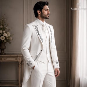May include: A white suit featuring a long jacket with black floral embroidery on the lapels and cuffs, a matching vest, a white dress shirt, a white necktie, and white trousers. The text SuitCrafters is visible in the upper right corner.