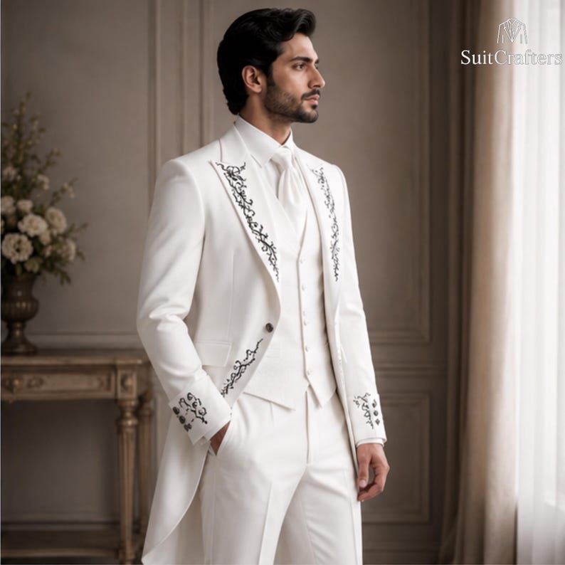 May include: A white suit featuring a long jacket with black floral embroidery on the lapels and cuffs, a matching vest, a white dress shirt, a white necktie, and white trousers. The text SuitCrafters is visible in the upper right corner.