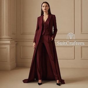 May include: A deep burgundy three-piece suit featuring a long, floor-length coat, a tailored vest with buttons, and matching trousers. The text SuitCrafters is visible on the right side. The ensemble is a solid, dark red color throughout.