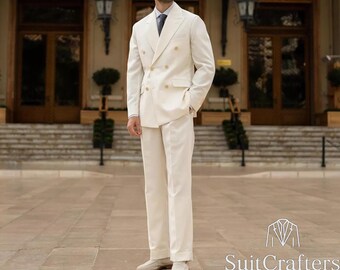 May include: A cream-colored double-breasted suit with gold buttons. The suit includes a jacket and matching trousers. The person is wearing a light blue tie and sunglasses. The logo "SuitCrafters" is visible in the bottom right corner.
