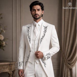 May include: A white suit featuring a long jacket with black floral embroidery along the lapels and cuffs, a matching white vest, and a white cravat. The jacket includes a single button closure. The text SuitCrafters is visible in the top right corner.