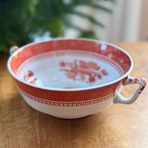 May include: A white and red ceramic bowl with two handles. The bowl has a red patterned rim and a red design inside. The bowl is sitting on a wooden surface. A green tree is in the background.