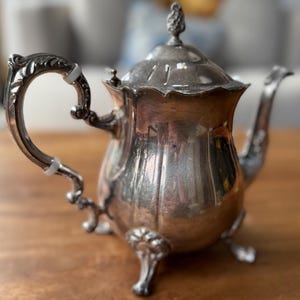 May include: A small, antique silver teapot with a curved handle and a decorative spout. The teapot has a lid with a small, ornate finial. It sits on three small feet. The teapot is displayed on a wooden surface.