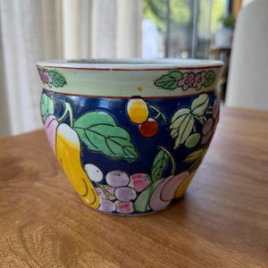 Puede incluir: Una maceta de cerámica redonda con un fondo azul intenso decorado con un patrón colorido de varias frutas, incluyendo peras amarillas, uvas moradas y cerezas rojas, rodeadas de hojas verdes y un borde verde claro con detalles florales.