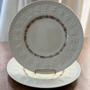 May include: Two off-white ceramic plates with embossed floral patterns and a decorative border. The plates have a colorful floral design around the inner rim. One plate is resting on a gold-colored plate stand.