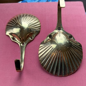 May include: Two brass seashell-shaped wall hooks. The hooks have a detailed, ridged design and a curved hook for hanging items. The hooks are mounted on a pink surface. The hooks are made of brass.