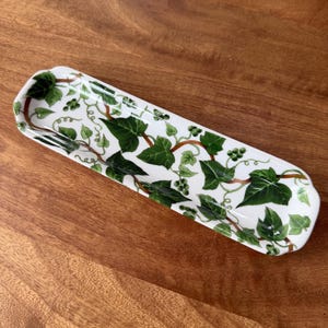 May include: A rectangular white ceramic serving dish with a green ivy leaf and vine pattern. The dish is approximately 12 inches long and 4 inches wide, with rounded corners. The ivy design features green leaves and brown vines.
