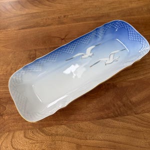 May include: A rectangular ceramic serving dish with a blue and white sky design and two seagulls in flight. The dish has a scalloped edge and a gold trim. The dish is approximately 12 inches long.