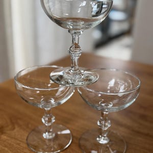 May include: Three clear glass coupe glasses arranged in a pyramid stack. Each glass features a wide, shallow bowl, a slender stem with a decorative central knob, and a circular base. The glassware is transparent and smooth with a polished finish.