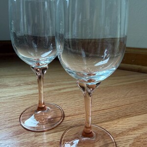May include: Two clear glass wine glasses featuring tall, transparent bowls and stems with a soft pink tint. Each glass has a wide, circular base that matches the pink color of the stem, providing a stable foundation for the upright drinking vessel.